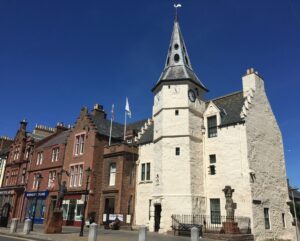 A row of red stone buildings connected to a building with white harling and dark grey roof tiles. At the front of the white building is a clocktower with a tall, pointed roof.