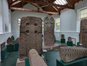 A collection of ornately carved Pictish stones displayed in a room with white walls and wooden beamed ceiling.