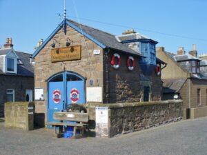 A small two-storey building with brown stone walls, pale blue trim, and a grey slate roof. Three red and white life rings hang from one wall. A wooden sign with the text "The Maggie Law Maritime Museum" hands above a large pale blue double door.