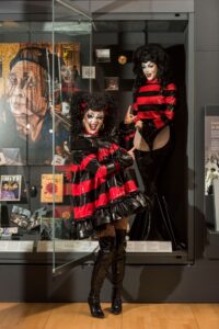 Ellie Diamond, a drag queen with a curly black wig and red and black vinyl dress, poses in front of a museum case. Inside the case is a mannequin with Ellie Diamond's face, a black curly wig, and a red and black vinyl bodysuit.