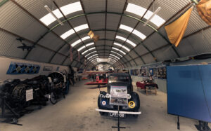 A long hangar with an arched metal roof. The hangar contains a black vintage car, a red biplane, and a variety of objects and displays relating to the history of flight.
