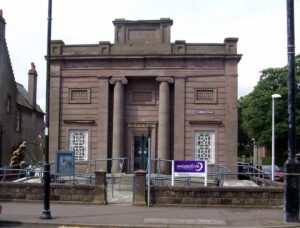A neo-classical building with ornately carved red stone walls. A doorway is situated between two tall pillars in the middle of the building.