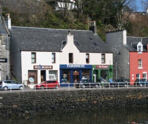 Image of Mull museum, next to two shop fronts. There are cars parked outside of the building and a large stone wall below with a white railing at the top.