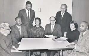 A black and white photograph depicting a group of people of mixed ethnicity, ages and gender sat around a table. The photograph captures the first meeting of the International Afro-American Museum (IAM) in 1965.