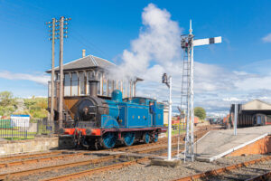 A bright blue and black steam locomotive billows smoke as it runs on a train track past a small signal box and raised platform.