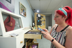 A smiling adult with bright red hair and light skin stands side on to the camera in front of a display of objects. They are holding a mobile phone.
