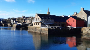 Photo of a harbour on a sunny day with a large grey building in the centre, a red shed-type building to its right and further smaller buildings to its left. The water is in the foreground.