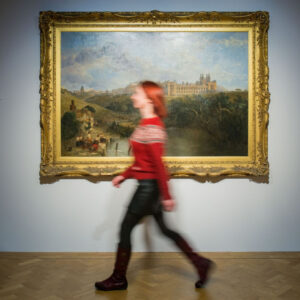 An adult with light skin, red hair, and a red jumper walks in front of a large painting of a stately home at the City Art Centre. The walking motion of the adult has caused their image to blur.