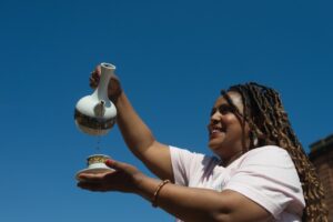 A smiling adult with medium-dark skin pours coffee from coffee pot into a cup. They are standing outdoors against a bright blue sky.