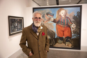 Artist John Byrne - a man with a white hair and beard and round glasses - stands in front of one of his paintings within an art gallery.