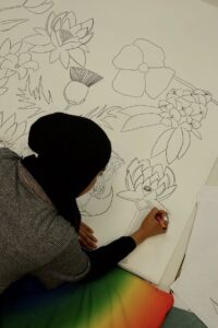 A person with medium skin tone wearing a black head scarf and striped top is leaning over a large sheet of paper full of hand drawn flowers. They are drawing a flower.