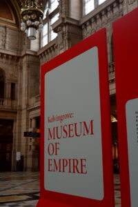 A large red and white sign in front of a highly decorated stone interior of a building. Sign text " Kelvingrove: Museum of Empire".