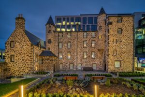 A 16th century stone house with circular stair towers with a planted garden and courtyard. The house is situated in a metropolitan area with modern glass buildings visible around it.