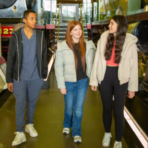 A teenager with medium-light skin and long brown hair, a teenager with medium-dark skin and short brown hair, and a teenager with light skin and long red hair walk through a display of steam locomotives at Riverside Museum.