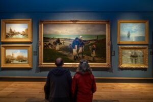 Two adults in raincoats sit on a bench in an art gallery with their backs to the camera. On the wall in front of them is a variety of landscape paintings.