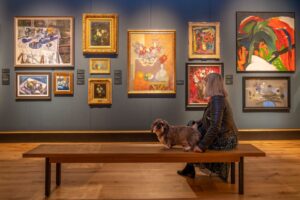 An adult with grey shoulder-length hair sits next to a small brown dog on a bench in an art gallery. On the wall behind them is a variety of colourful still-life paintings.