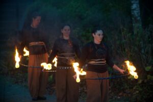 Three adults with light skin, brown jumpers and loose brown trousers walk in a line outside in the dark. Each adult holds flaming torches and a large ring around their waists.