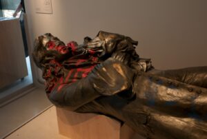 A grey metal statue of a man with long curly hair and wearing a frock coat is placed lying down on a wooden plinth in a room. The statue has red and blue spray paint on the face and body.