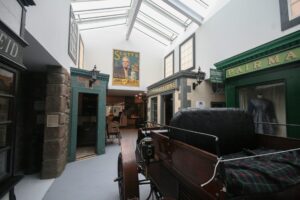 A long museum gallery with a glass ceiling. Shop fronts on either side of the gallery evoke a 19th century Scottish high street. In the middle of the gallery is a wooden open-top carriage with leather seats.