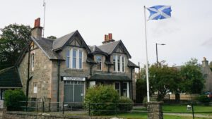 A two-storey building with beige stone walls, a grey tiled roof, and white windows. A sign above a window on the ground floor reads "Clan Macpherson House & Museum". The flag of Scotland flies on a flagpole in front of the building.