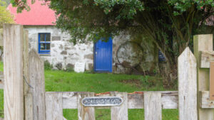 A small one-storey stone building with a red metal roof and a blue wooden door. In front of the building is a tree with low branches and a wooden gate with a small sign which displays the text "please shut the gate".