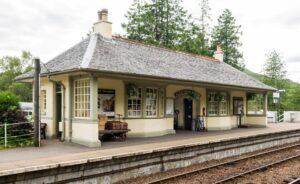 A single-storey building on a railway platform. It has a grey tiled roof, cream walls, and pale green accents. The building and platform are decorated with 20th century railway posters and station equipment.