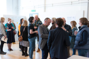 Several small groups of adults stand and engage in conversation in a room with white walls and a light wooden floor.