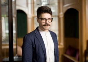 An adult with light skin, short brown hair, and a brown moustache. They have glasses, a nose ring, a white shirt, and a dark blue blazer.