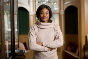 An adult with medium-dark skin and long dark-brown hair which has been tied back behind their head. They are wearing a beige turtleneck jumper.