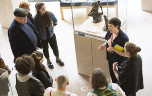 A group of people are standing together, listening to a tour guide speaking. The group of people is made up of a mix of younger and older people. The tour guide has a light skin tone and short brown hair.