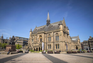 A two-storey building with walls of warm brown stone and a grey tiled roof. It has gothic features which include small turrets and arched windows, and located in the centre of a small civic square.