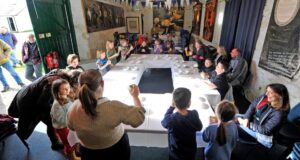 A group of adults and children sit around a white table in a small room with rough white stone walls. The children are shaking glass jars filled with liquid.