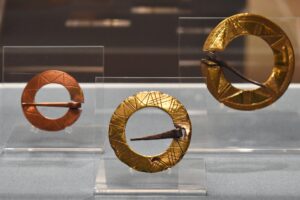 Three circular metal brooches displayed in a row. Two of the brooches have a gold colour, while the other has a copper colour. All three brooches are decorated with linear incisions.