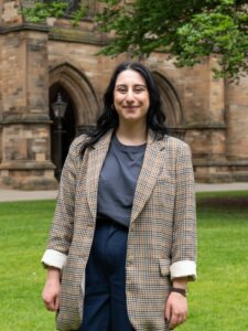 An adult with light skin, long black wavy hair, and a brown check jacket. They are standing on a lawn in a courtyard.