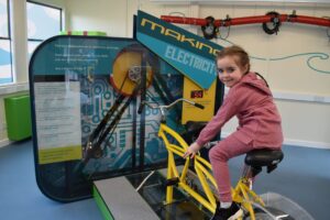 A child with light skin and long braided brown hair sits on a yellow tandem bike. The bike is attached to the floor and an interactive panel which reads "Making Electricity".