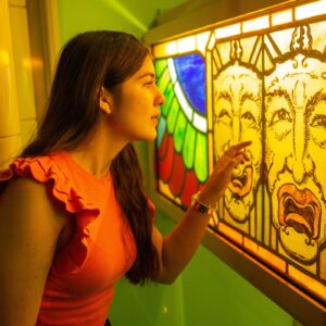 A teenager with medium-light skin and long brown hair raises their hand up to a stained glass panel. The panel is decorated with two detailed faces.