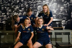 Five teenagers in football uniforms laugh and pose on a bench. On the wall behind them is a black-and-white photograph of a crowd.