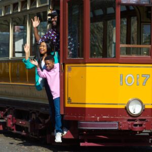 An adult with dark skin, an adult with medium dark skin, and a child with medium-dark skin laugh and wave as they lean out of the side of an old red and yellow tram.
