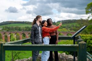 Three adults pose for a selfie in front of rolling hills, green trees, and a long red stone viaduct. The adult on the left has light skin, brown hair in a ponytail, and a beard. The adult in the middle has medium-tone skin and shoulder length dark brown hair. The adult on the right has dark skin, glasses, and long dark brown braided hair.