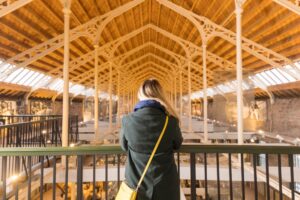 An adult with long blonde hair and their back turned to the camera looks out from a first-floor balcony inside a long hall. The hall has a pointed wooden roof supported by slender white metal pillars.