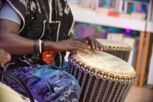 An adult with dark skin and a patterned black tunic uses their hands to play a drum.