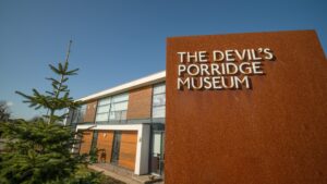 A slab of rusted metal which displays the words "The Devil's Porridge Museum". Behind the sign is a pine tree and a two-storey building with large windows and wood panel walls.