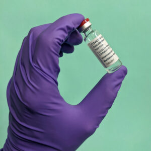 A hand, wearing a purple surgical glove, holds up a glass COVID-19 vaccine vial.