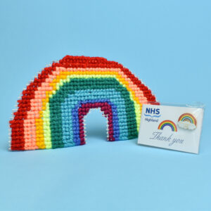 A woven rainbow decoration displayed next to a small white card. A rainbow pin badge is attached to the card. Printed onto the card are the NHS Highland logo, a rainbow, and the text "Thank you".