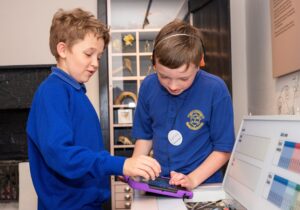 Two children with light skin, medium-length brown hair, and bright blue school uniforms interact with a digital tablet. One of the children wears headphones which are connected to the device.