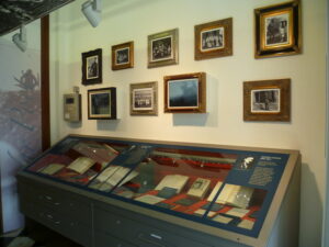 Two lines of wooden framed, black and white photographs hang on a wall above a glass topped display case. The glass case contains journals and books, open for people to read their contents.