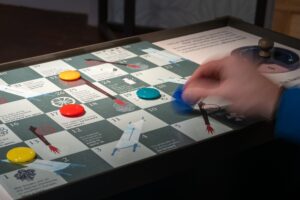 A person plays an educational board game on the topic of battlefield hospitals in the style of snakes and ladders. A hand, blurred with motion, holds a blue counter over the board.