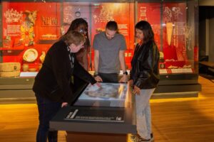 Three young adults with medium skin tone and brown hair, light skin tone and brown hair, and dark skin tone, glasses, and braided hair are stood looking down at a digital display with an image of a map on it. An older adult with light skin tone, light brown hair, wearing glasses and a lanyard is pointing at the display. Behind them is a floor to ceiling glass display case including animal skulls, domestic pottery items and interpretation text and images.