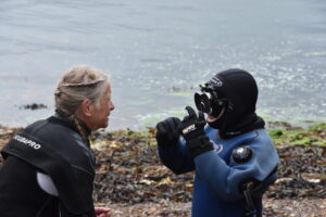 Two people in wet suits on the sea shore. One explaining to the other how to use snorkel mask.