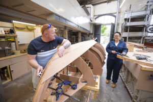 Two adults holding tools working on the frame of an upturned wooden boat. One adult has light skin and short red hair; the other has light skin and blonder hair tied into a bun.